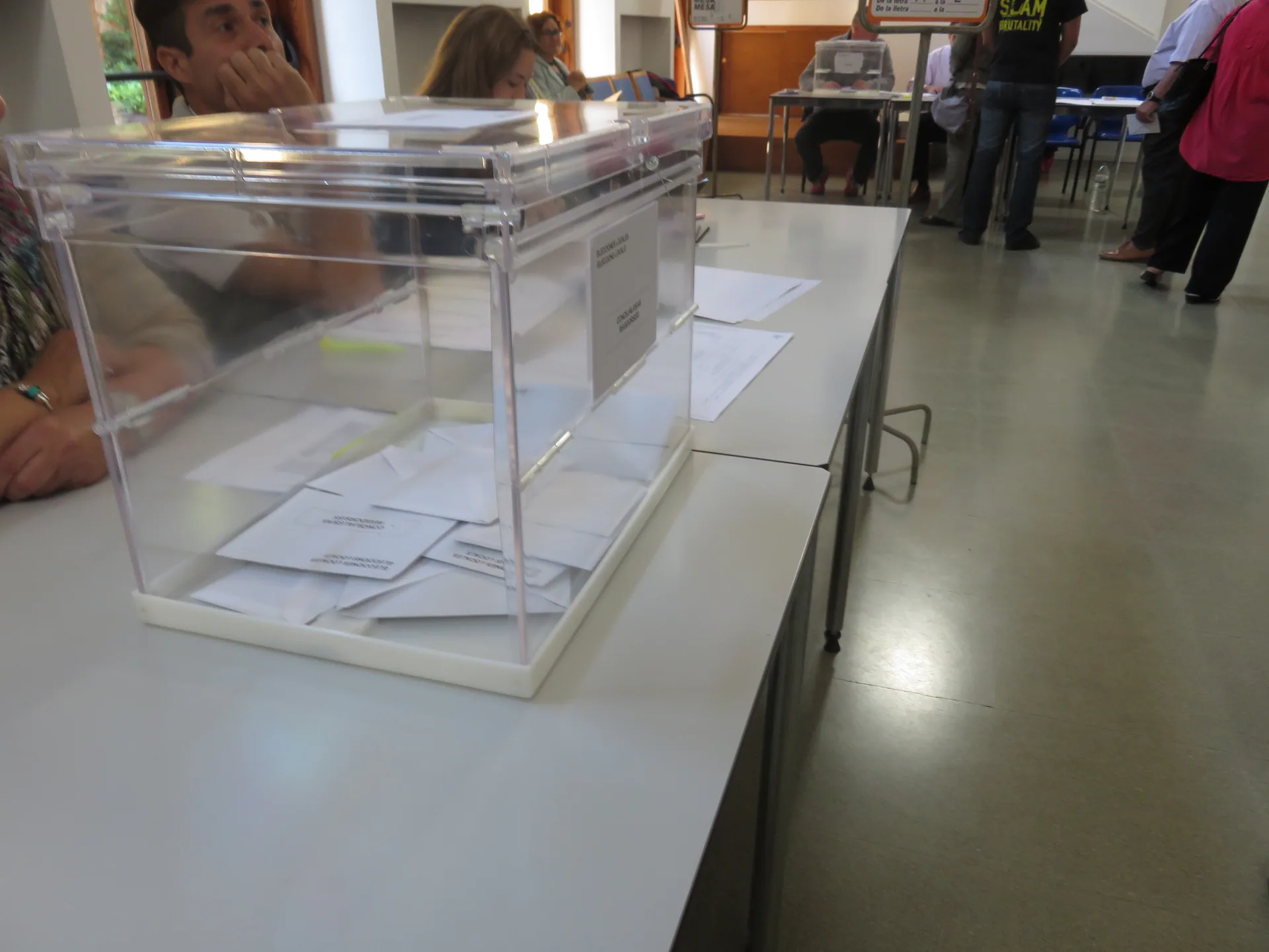 Una urna transparente con papeletas sobre la mesa electoral, con los miembros de la mesa sentados al lado
