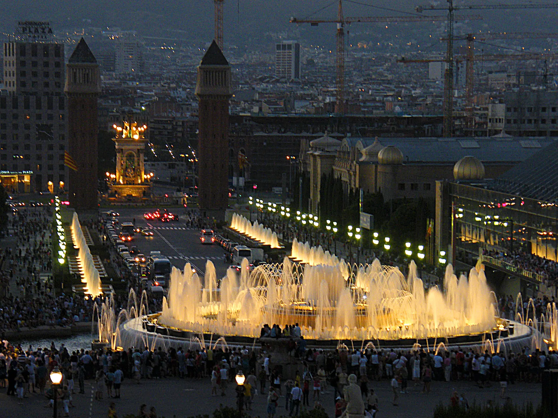 The Magic Fountain of Montjuïc lit up at night, with crowds around it and the Plaça d'Espanya towers behind