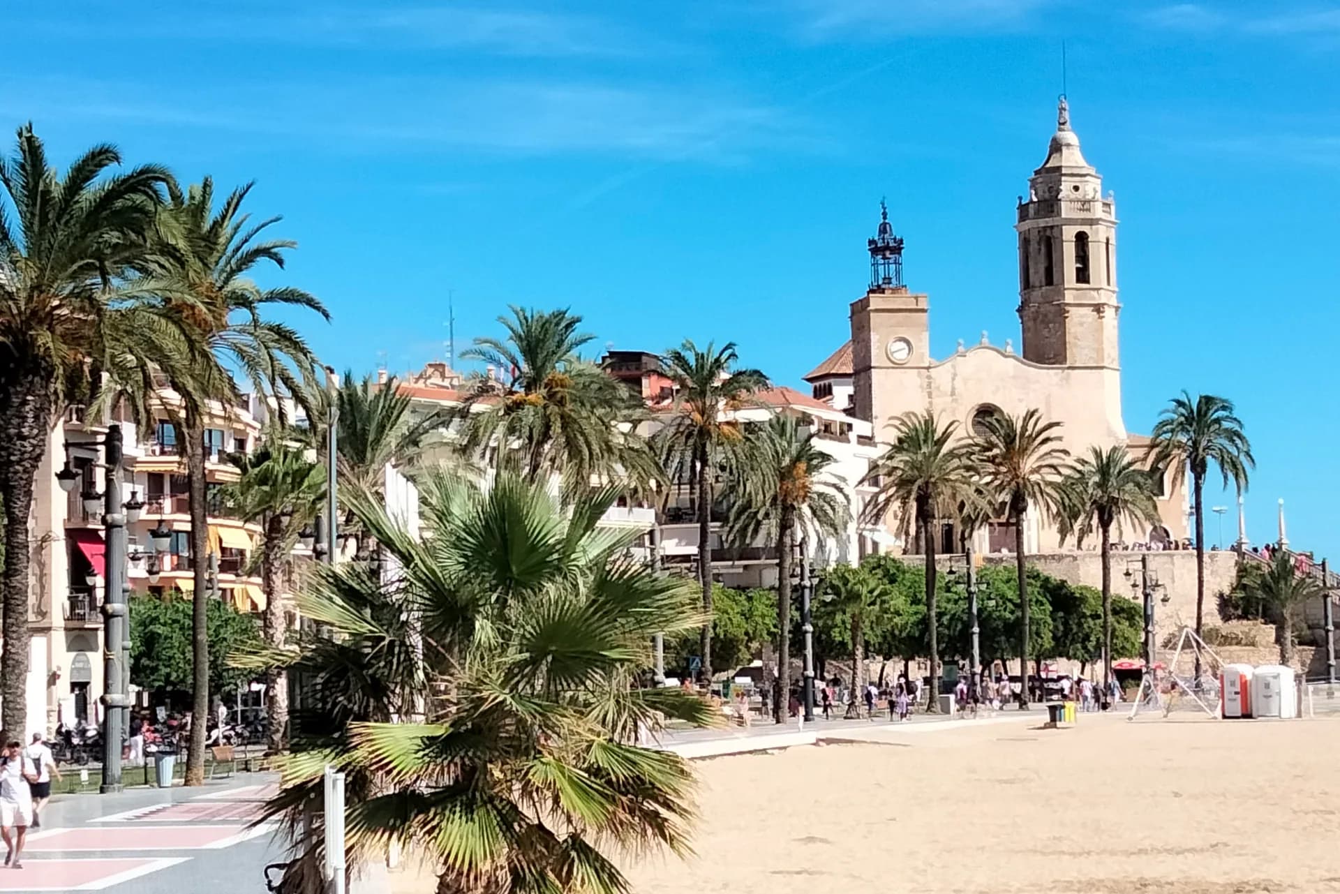 La playa de la Fragata de Sitges con palmeras y la iglesia de Sant Bartomeu i Santa Tecla al fondo